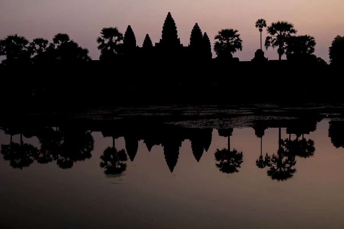 Angkor Wat towers in silhouette at sunrise with palms and pond reflection