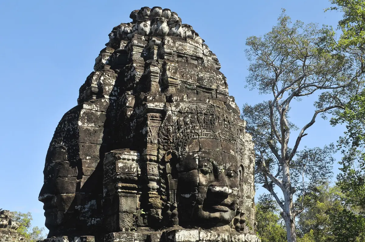 Side view of a Bayon tower with a carved face and leafy trees behind